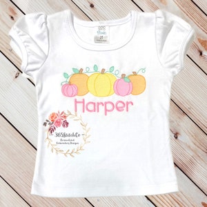 May include: White short-sleeved shirt with a vine of pumpkins and the name "Harper" embroidered on the front. The shirt is decorated with a floral wreath and the text "365StitchCo - Personalized Embroidery Designs".
