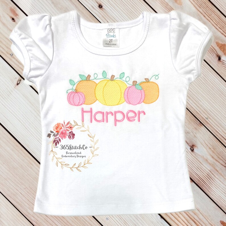 May include: White short-sleeved shirt with a vine of pumpkins and the name "Harper" embroidered on the front. The shirt is decorated with a floral wreath and the text "365StitchCo - Personalized Embroidery Designs".