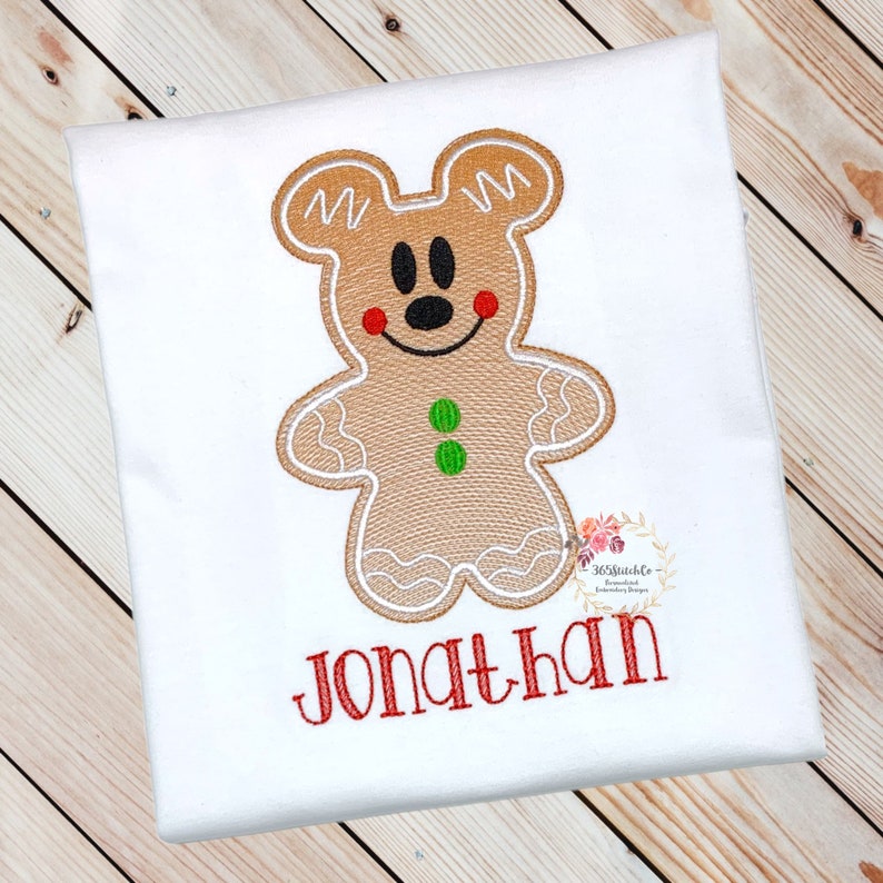 May include: A white t-shirt with an embroidered gingerbread man design. The gingerbread man is brown with white icing and has a red nose and green buttons. The name "Jonathan" is embroidered in red below the gingerbread man.