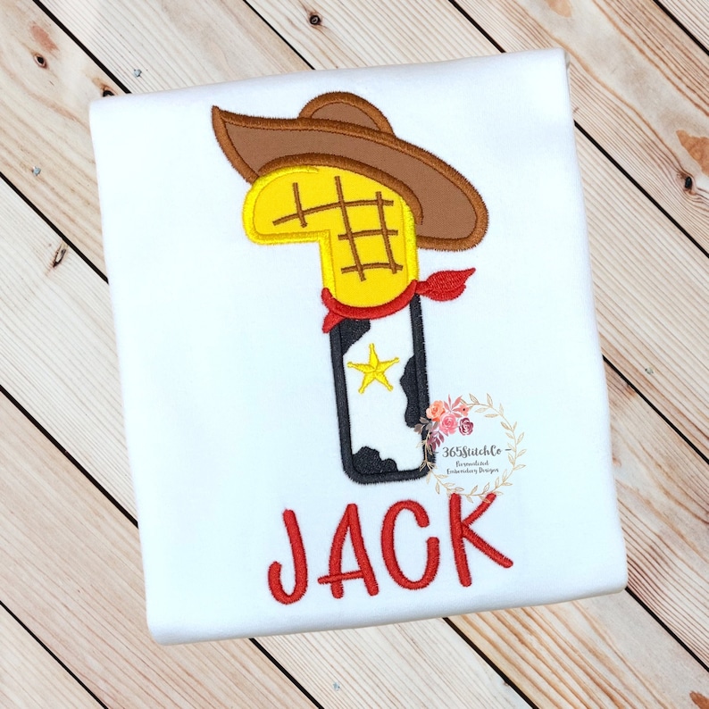 K&ouml;nnte beinhalten: Ein wei&szlig;er Strampler mit einem roten gestickten Cowboy-Stiefel mit einem gelben Stern, einem braunen Cowboyhut und einem roten Bandana. Der Strampler hat auch den Namen "JACK" in Rot gestickt.