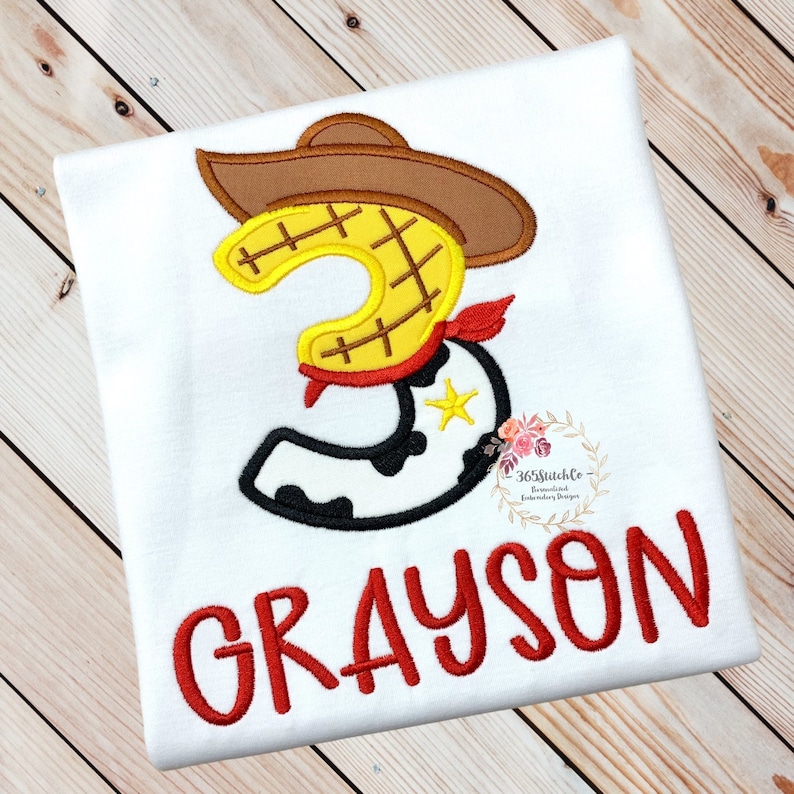 K&ouml;nnte beinhalten: Wei&szlig;es T-Shirt mit einer roten gestickten Nummer drei in Form eines Cowboy-Stiefels mit einem braunen Hut und einem roten Bandana. Der Name Grayson ist in rot unter der Nummer gestickt.