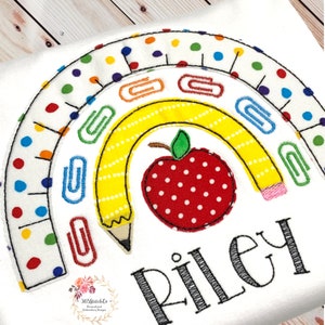 May include: A rainbow-shaped design with a yellow pencil, red apple, and paper clips. The rainbow is made of white fabric with colourful polka dots. The text "Riley" is embroidered below the design.