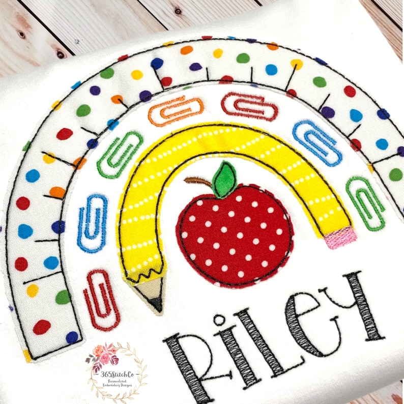 May include: A rainbow-shaped design with a yellow pencil, red apple, and paper clips. The rainbow is made of white fabric with colourful polka dots. The text "Riley" is embroidered below the design.