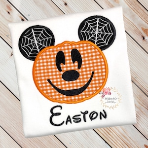 K&ouml;nnte beinhalten: Wei&szlig;es T-Shirt mit einem gestickten K&uuml;rbis-Mickey-Mouse-Design. Der K&uuml;rbis ist orange und wei&szlig; kariert mit einem schwarzen gestickten L&auml;cheln und Augen. Die Ohren sind schwarz mit wei&szlig;en Spinnweben. Der Name Easton ist unter dem K&uuml;rbis-Mickey-Mouse gestickt.