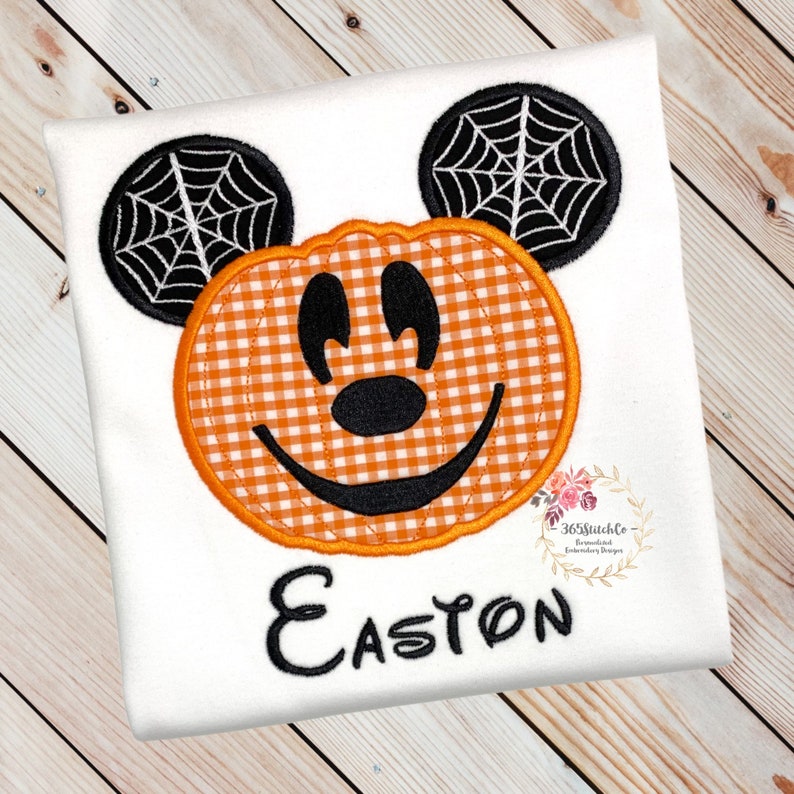K&ouml;nnte beinhalten: Wei&szlig;es T-Shirt mit einem gestickten K&uuml;rbis-Mickey-Mouse-Design. Der K&uuml;rbis ist orange und wei&szlig; kariert mit einem schwarzen gestickten L&auml;cheln und Augen. Die Ohren sind schwarz mit wei&szlig;en Spinnweben. Der Name Easton ist unter dem K&uuml;rbis-Mickey-Mouse gestickt.