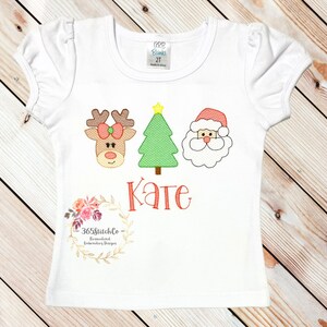 Puede incluir: Camiseta blanca con un reno rojo, un &aacute;rbol de Navidad verde y un Pap&aacute; Noel bordados. El nombre "Kate" est&aacute; bordado debajo de los personajes. La camiseta tiene una peque&ntilde;a etiqueta que dice "365Stitchlo -  Personalized Embroidery Designs".