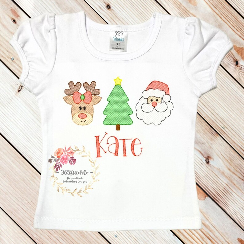 Puede incluir: Camiseta blanca con un reno rojo, un &aacute;rbol de Navidad verde y un Pap&aacute; Noel bordados. El nombre "Kate" est&aacute; bordado debajo de los personajes. La camiseta tiene una peque&ntilde;a etiqueta que dice "365Stitchlo -  Personalized Embroidery Designs".
