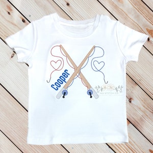 Op de afbeelding: Wit T-shirt met geborduurde hengels, rood en blauw touw en harten. De naam "Cooper" is geborduurd op het shirt.