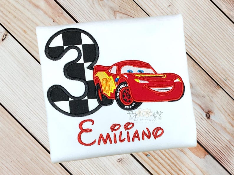 Pu&ograve; includere: T-shirt bianca con una macchina da corsa rossa e il numero 3 con un motivo a bandiera a scacchi. Il nome "Emiliano" &egrave; ricamato in rosso sotto. Un design divertente per il compleanno di un bambino.