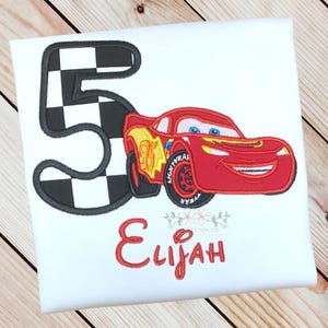 Pu&ograve; includere: T-shirt bianca con un'auto da corsa rossa e il numero 5 con un motivo a bandiera a scacchi. Il nome "Elijah" &egrave; ricamato in rosso. Un design divertente per il compleanno di un bambino.