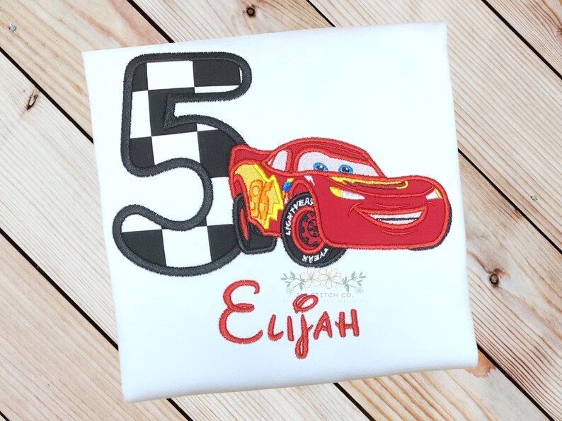Pu&ograve; includere: T-shirt bianca con un'auto da corsa rossa e il numero 5 con un motivo a bandiera a scacchi. Il nome "Elijah" &egrave; ricamato in rosso. Un design divertente per il compleanno di un bambino.