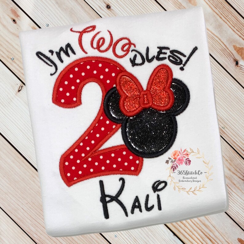 May include: A white t-shirt with red and black embroidery. The embroidery includes the text "I'm Two-dles!" and a red number "2" with white polka dots. A black Mickey Mouse head with a red bow is next to the number. The name "Kali" is embroidered below the number.