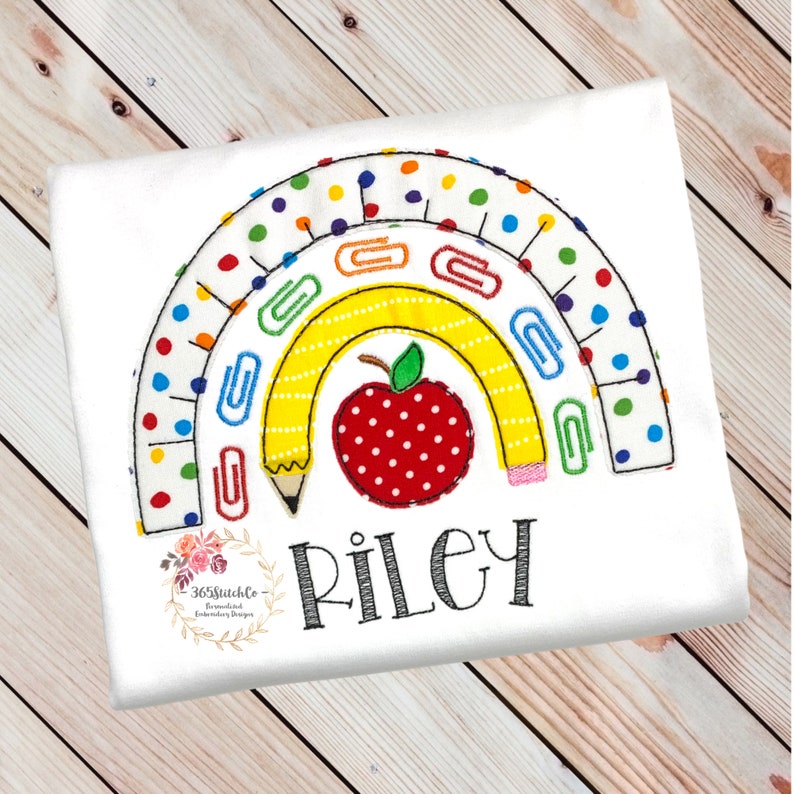 May include: A white t-shirt with an embroidered rainbow design featuring a yellow pencil, red apple, and paper clips. The name "Riley" is embroidered below the rainbow.