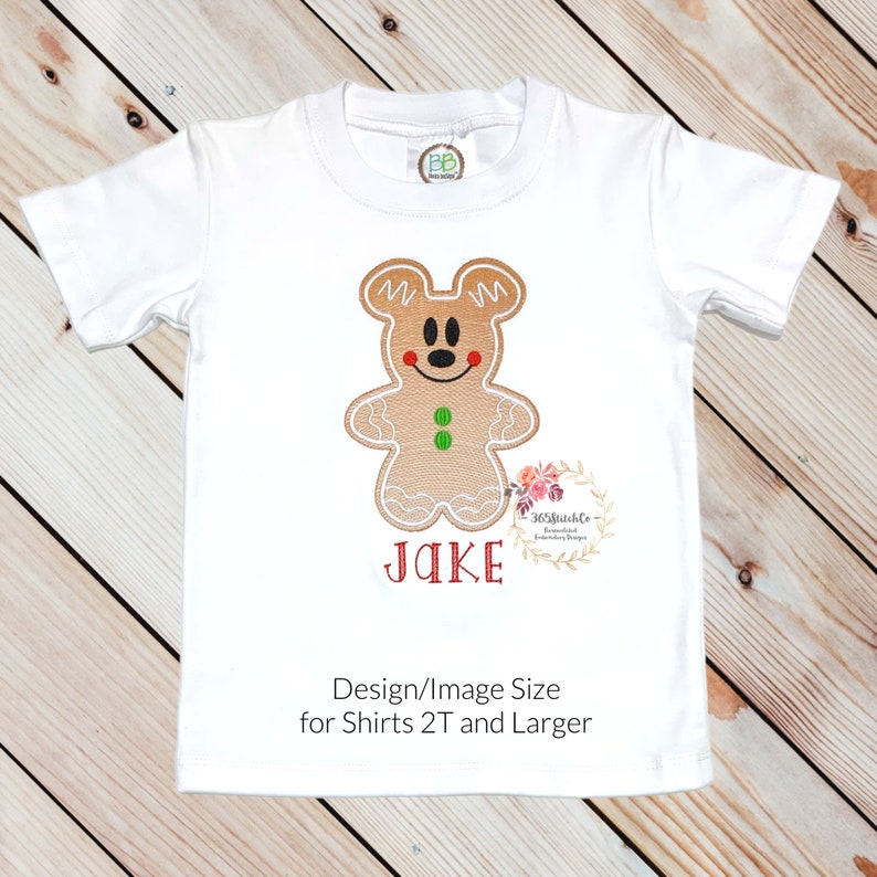 May include: White t-shirt with an embroidered gingerbread man design. The gingerbread man is brown with red and green accents. The name "JAKE" is embroidered in red below the gingerbread man. The text "Design/Image Size for Shirts 2T and Larger" is printed in black at the bottom of the shirt.