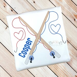 Op de afbeelding: Geborduurde afbeelding van twee gekruiste hengels met rood en blauw touw, een hartvorm aan het einde van elk touw en de naam "Cooper" in blauwe letters.
