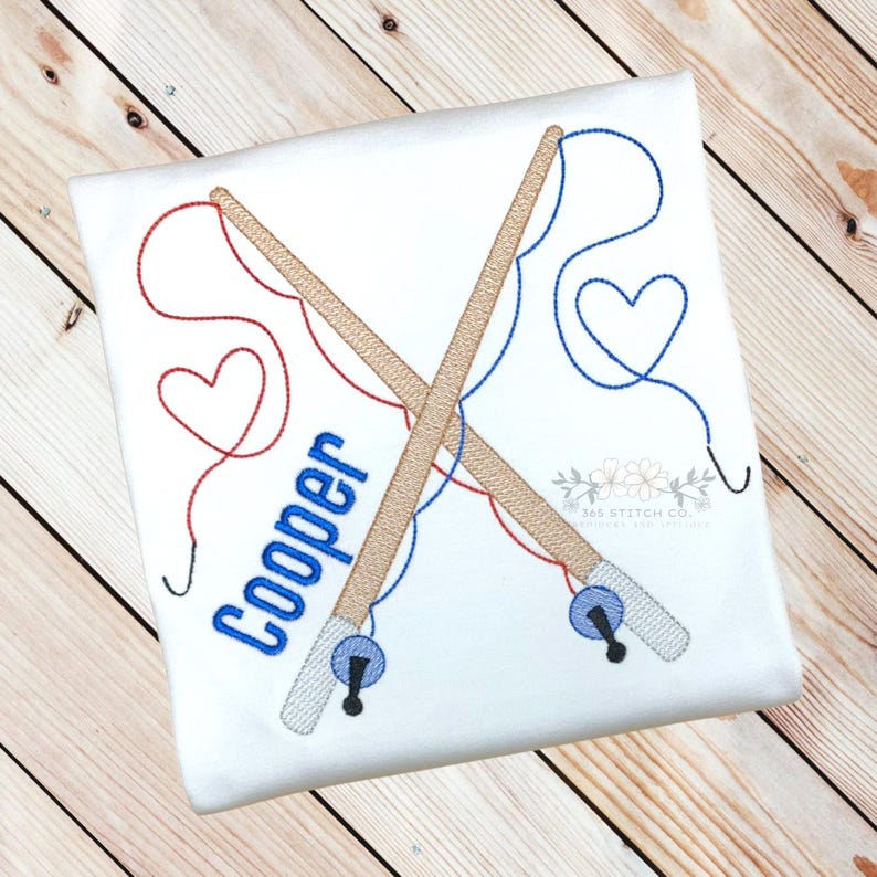 Op de afbeelding: Geborduurde afbeelding van twee gekruiste hengels met rood en blauw touw, een hartvorm aan het einde van elk touw en de naam "Cooper" in blauwe letters.