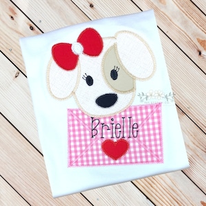 May include: A white and tan dog with a red bow is peeking out of a pink and white gingham envelope with a red heart. The envelope says "Brielle" in black.