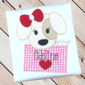 May include: White t-shirt featuring an embroidered applique of a dog holding a pink and white gingham envelope with the name "Adeline" embroidered on it.