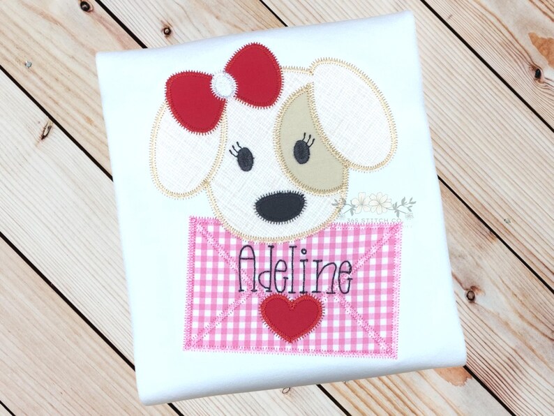 May include: White t-shirt featuring an embroidered applique of a dog holding a pink and white gingham envelope with the name "Adeline" embroidered on it.