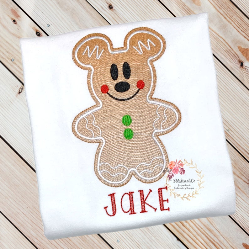 May include: A white t-shirt with an embroidered gingerbread man design. The gingerbread man is brown with white icing and green buttons. The gingerbread man has a Mickey Mouse face. The shirt also has the name "JAKE" embroidered in red below the gingerbread man.