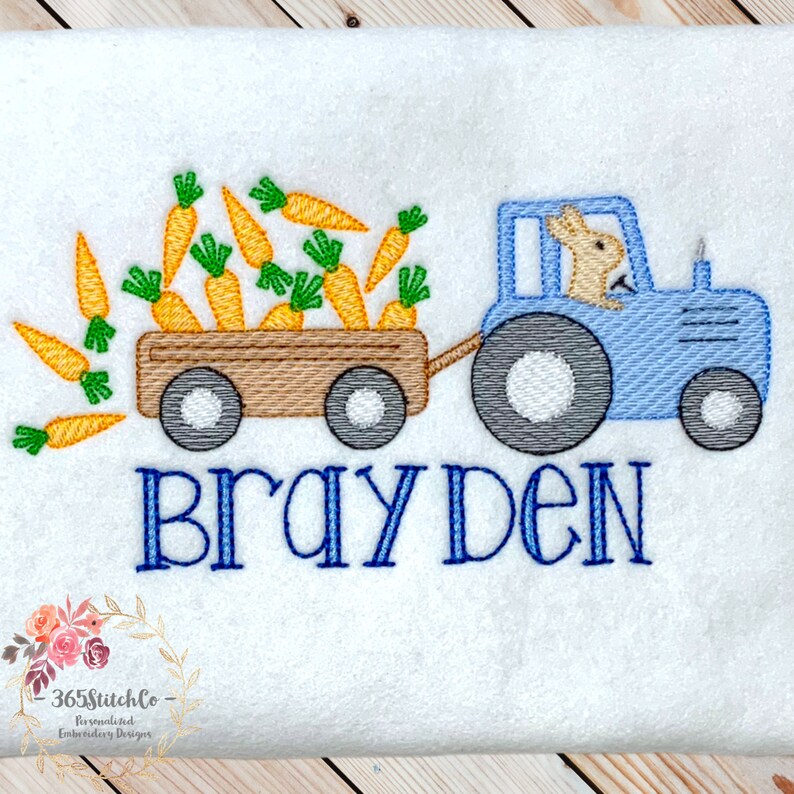 May include: A blue tractor with a bunny driver pulling a wagon full of orange carrots. The wagon has brown wheels and the tractor has gray wheels. The text "Brayden" is embroidered below the tractor.