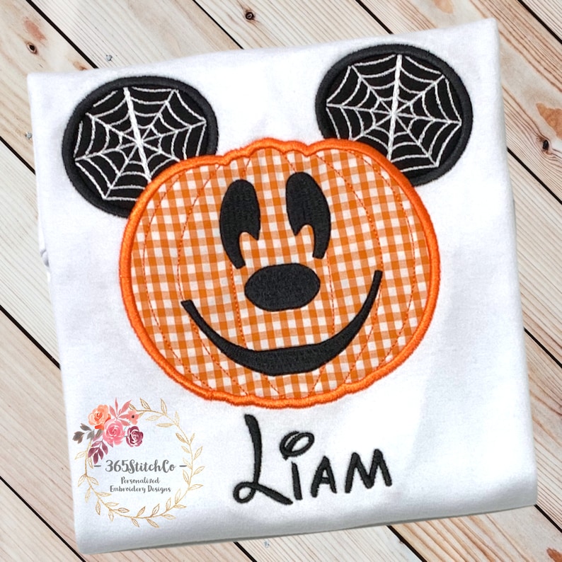 K&ouml;nnte beinhalten: Wei&szlig;es T-Shirt mit einem orange-schwarzen K&uuml;rbis-Mickey-Mouse-Design. Der K&uuml;rbis hat ein Karomuster und die Ohren haben ein schwarzes Spinnennetz-Design. Das Shirt tr&auml;gt auch den Namen "Liam" gestickt.
