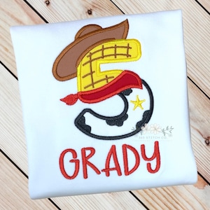 K&ouml;nnte beinhalten: Wei&szlig;es T-Shirt mit einer roten, gelben und braunen Stickerei der Nummer 5 in Form von Woody aus Toy Story. Das Shirt tr&auml;gt au&szlig;erdem den Namen "GRADY" in roter Stickerei.