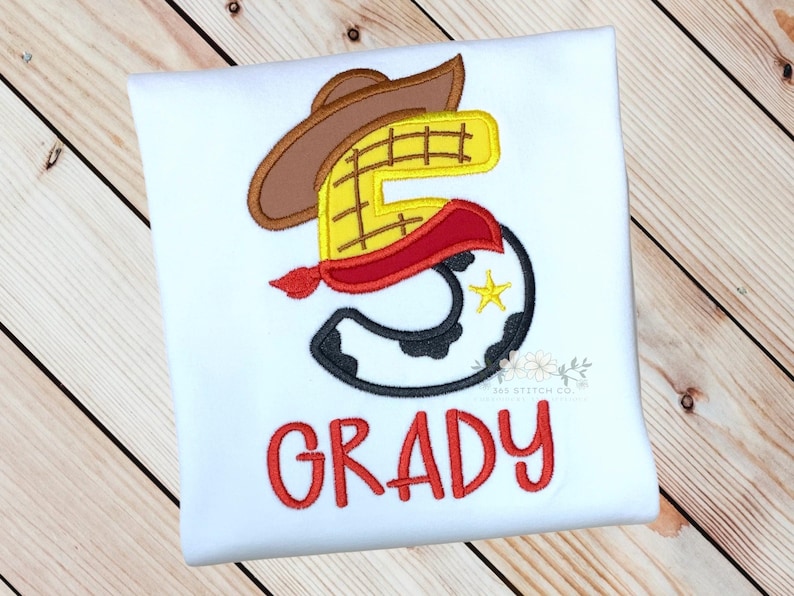 K&ouml;nnte beinhalten: Wei&szlig;es T-Shirt mit einer roten, gelben und braunen Stickerei der Nummer 5 in Form von Woody aus Toy Story. Das Shirt tr&auml;gt au&szlig;erdem den Namen "GRADY" in roter Stickerei.