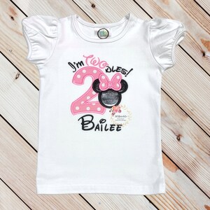 Puede incluir: Camiseta blanca de manga corta con bordado rosa y negro. El bordado dice "I'm Twodles!" con un n&uacute;mero 2 rosa y una cabeza de Minnie Mouse negra con un lazo rosa. El nombre "Bailee" est&aacute; bordado debajo del n&uacute;mero.