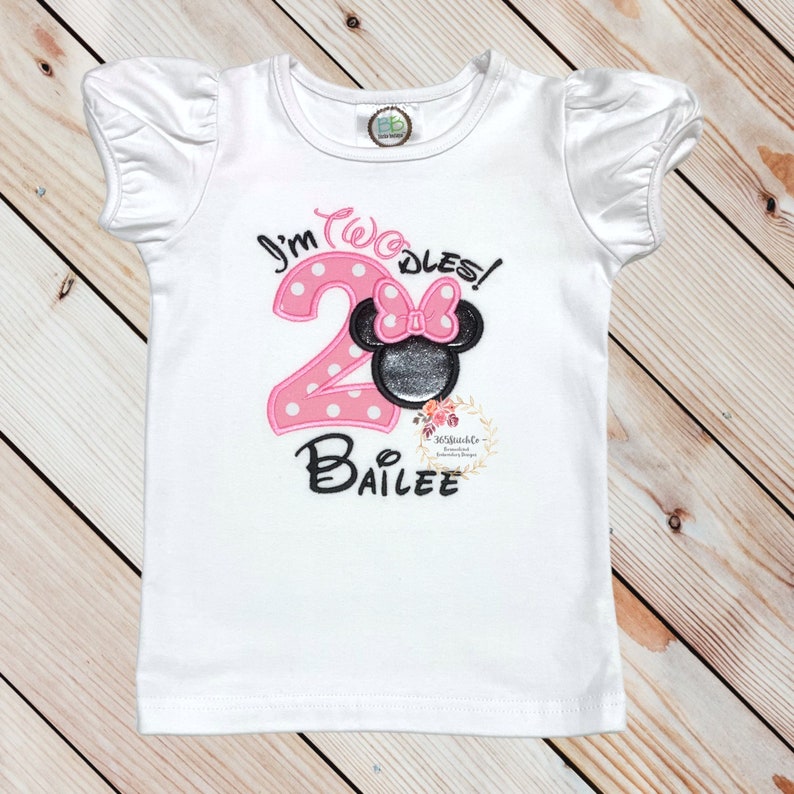 Puede incluir: Camiseta blanca de manga corta con bordado rosa y negro. El bordado dice "I'm Twodles!" con un n&uacute;mero 2 rosa y una cabeza de Minnie Mouse negra con un lazo rosa. El nombre "Bailee" est&aacute; bordado debajo del n&uacute;mero.