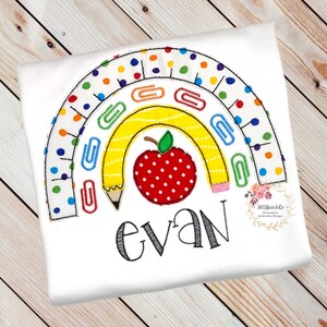 May include: A white t-shirt with an embroidered rainbow design featuring a yellow pencil, a red apple with white polka dots, and paper clips. The name "evan" is embroidered below the rainbow.