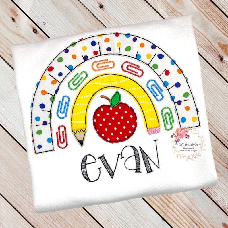 May include: A white t-shirt with an embroidered rainbow design featuring a yellow pencil, a red apple with white polka dots, and paper clips. The name "evan" is embroidered below the rainbow.