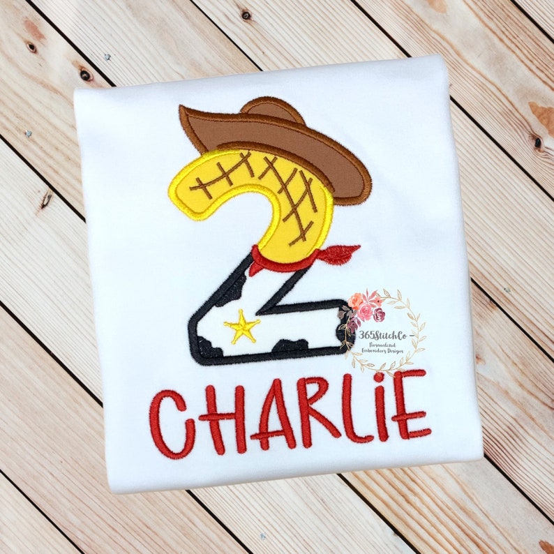 K&ouml;nnte beinhalten: Wei&szlig;es T-Shirt mit einer roten gestickten Zahl Zwei, einem Cowboyhut und einem roten Bandana. Die Zahl Zwei ist gelb mit einem schwarz-wei&szlig;en Kuhmuster unten. Der Text "CHARLIE" ist in Rot unter der Zahl gestickt.