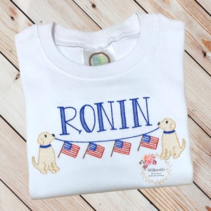Op de afbeelding: Een witte baby romper met de naam "RONIN" geborduurd in blauw. Twee gele honden houden een string met Amerikaanse vlaggen vast.