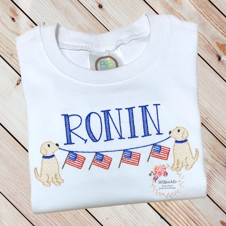 Op de afbeelding: Een witte baby romper met de naam "RONIN" geborduurd in blauw. Twee gele honden houden een string met Amerikaanse vlaggen vast.