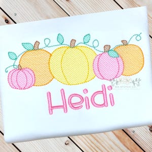 May include: A white onesie with an embroidered design of a row of yellow, orange, and pink pumpkins with green vines. The name "Heidi" is embroidered in pink below the pumpkins.