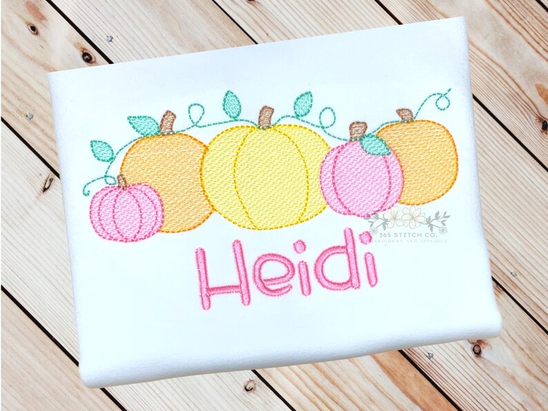 May include: A white onesie with an embroidered design of a row of yellow, orange, and pink pumpkins with green vines. The name "Heidi" is embroidered in pink below the pumpkins.