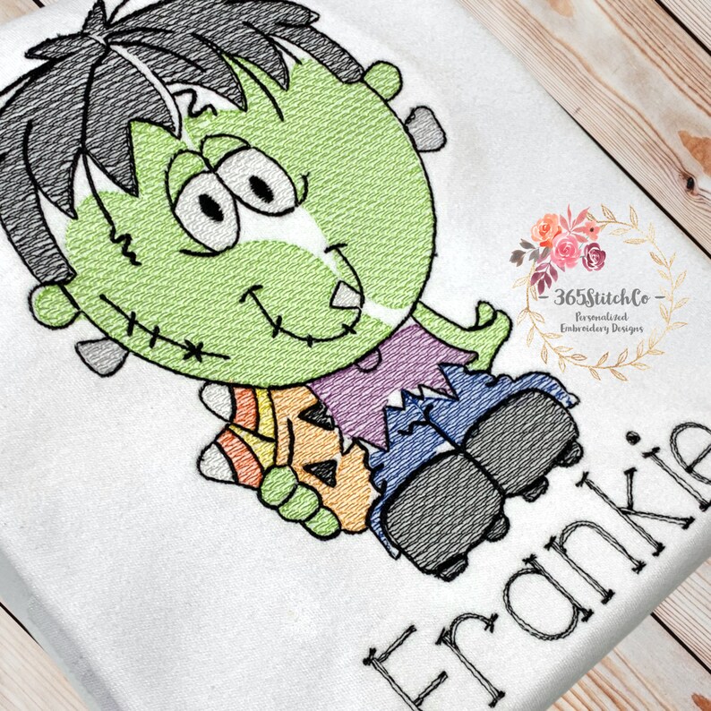 May include: Embroidered design of a green Frankenstein monster holding a candy corn and a pumpkin. The design is on a white fabric with the name Frankie embroidered below.