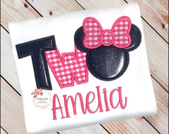May include: A white t-shirt with a black and pink "TWO" with a pink and white gingham pattern. A black and pink Minnie Mouse head is above the "TWO". The name "Amelia" is embroidered in pink below the "TWO".