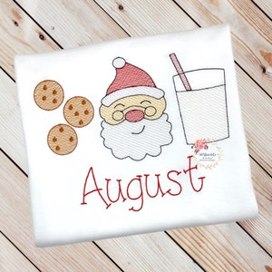 May include: Embroidered design featuring a cartoon Santa Claus with a glass of milk and cookies. The name "August" is stitched in red below the design.