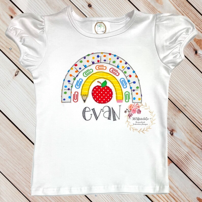 May include: White t-shirt featuring a rainbow design made from colourful pencils, paper clips and a red apple. The name "evan" is embroidered beneath the rainbow.