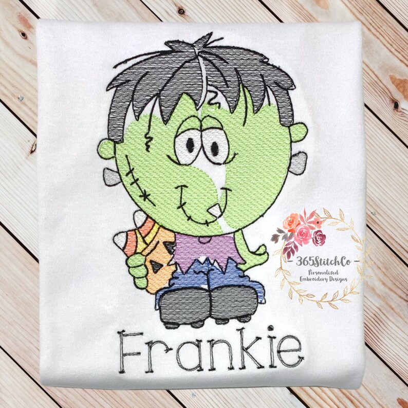 May include: Embroidered design of a green Frankenstein monster holding a candy corn. The monster has black hair, a purple shirt, blue jeans, and black shoes. The design includes the name "Frankie" in black lettering.