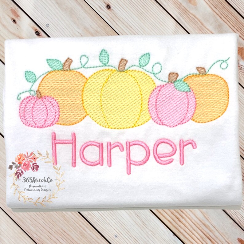 May include: A white t-shirt with an embroidered design of five pumpkins with green vines and leaves. The pumpkins are orange, yellow, and pink. The name "Harper" is embroidered in pink below the pumpkins.