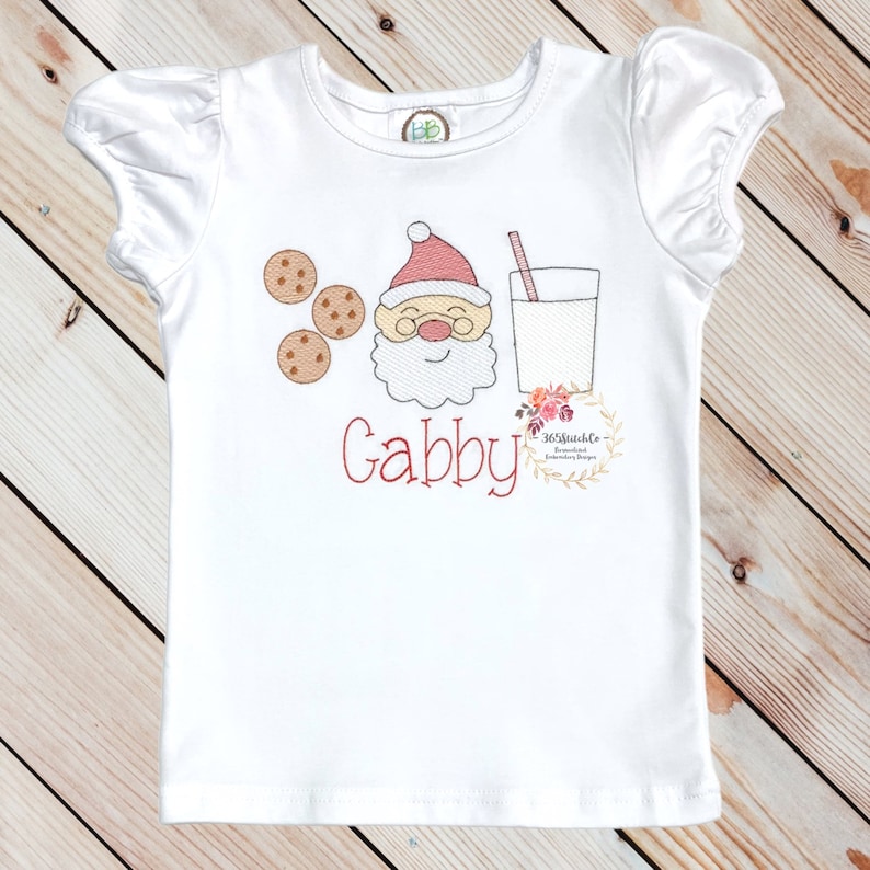 May include: White short-sleeved shirt with a red embroidered Santa Claus face, three cookies, and a glass of milk. The name "Gabby" is embroidered in red below the image.