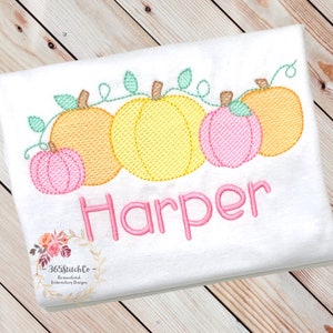 May include: Embroidered design of a row of five pumpkins with vines and leaves. The pumpkins are in shades of orange, yellow, and pink. The name "Harper" is embroidered below the pumpkins in pink thread.