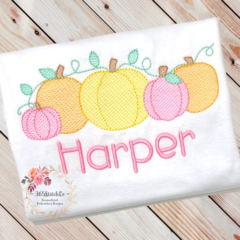 May include: Embroidered design of a row of five pumpkins with vines and leaves. The pumpkins are in shades of orange, yellow, and pink. The name "Harper" is embroidered below the pumpkins in pink thread.