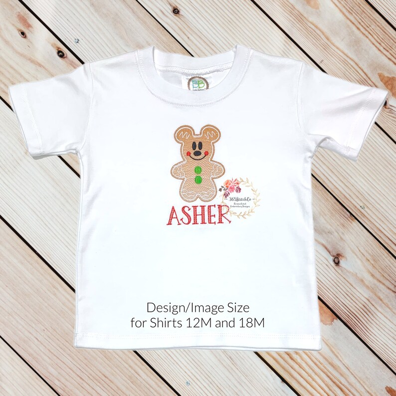 May include: White t-shirt with an embroidered gingerbread man design and the name "ASHER" in red. The design is suitable for shirts sized 12M and 18M.