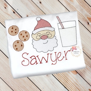 May include: A white embroidered shirt featuring a red Santa Claus face, three chocolate chip cookies, a glass of milk with a straw, and the name "Sawyer" in red embroidery.