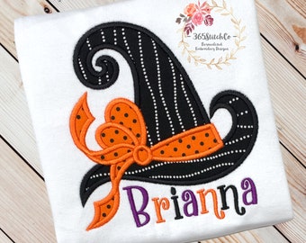 May include: A white t-shirt with an embroidered black and white witch hat with an orange and black polka dot bow. The name Brianna is embroidered in purple below the hat.