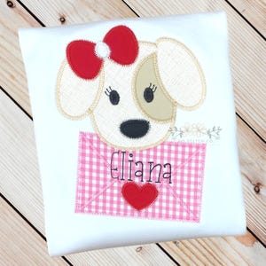 May include: White t-shirt with an embroidered applique of a dog holding a pink and white gingham envelope with a red heart and the name "Eliana" embroidered on it.